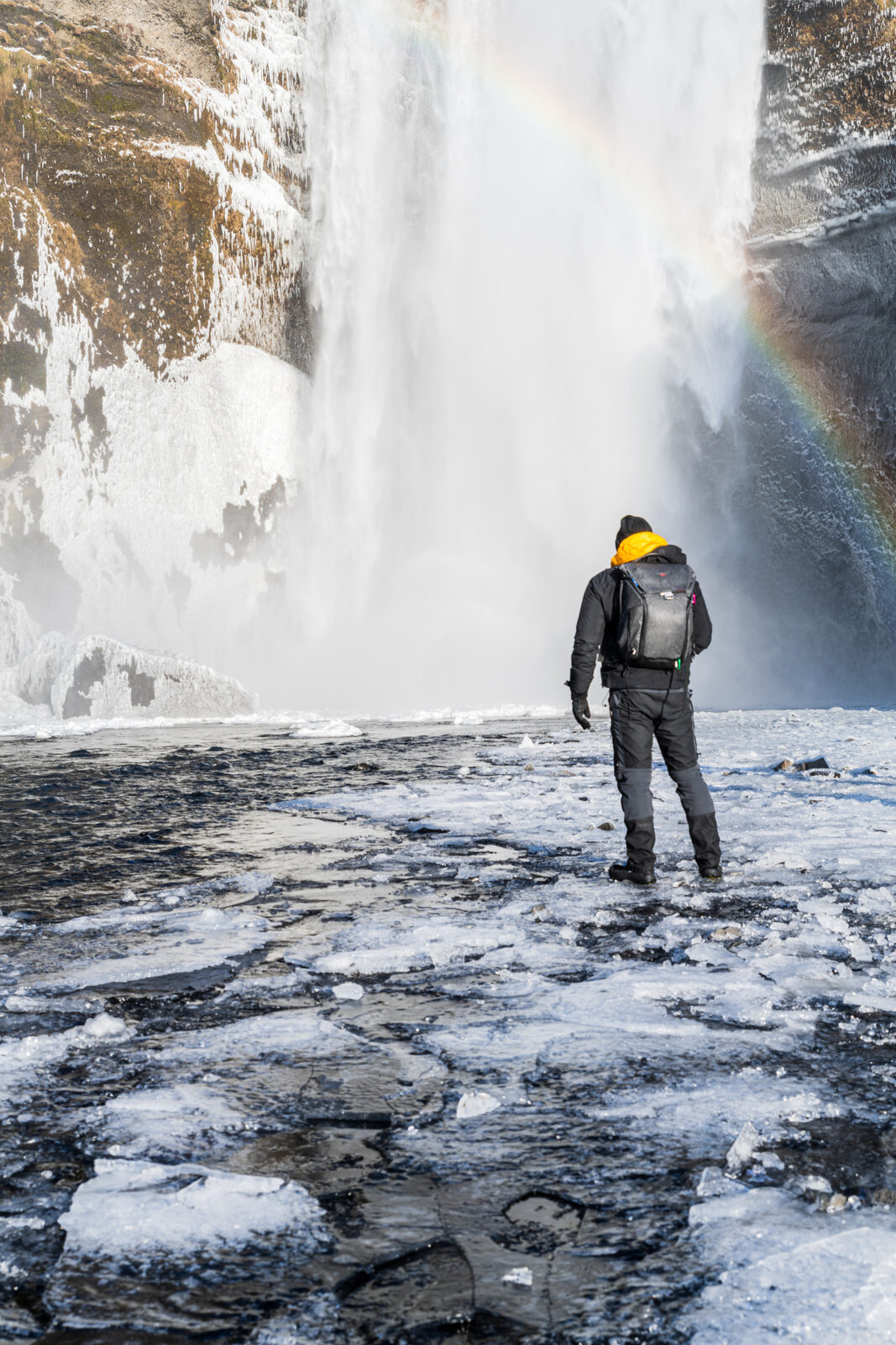 A hiker on the frozen riverbed below Skógafoss, a rainbow in the spray