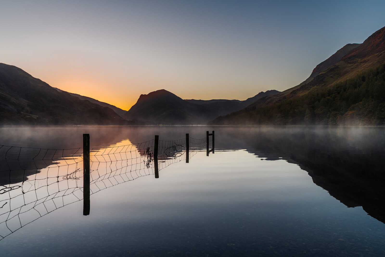 A fence running out into Buttermere at sunrise, mist on the water