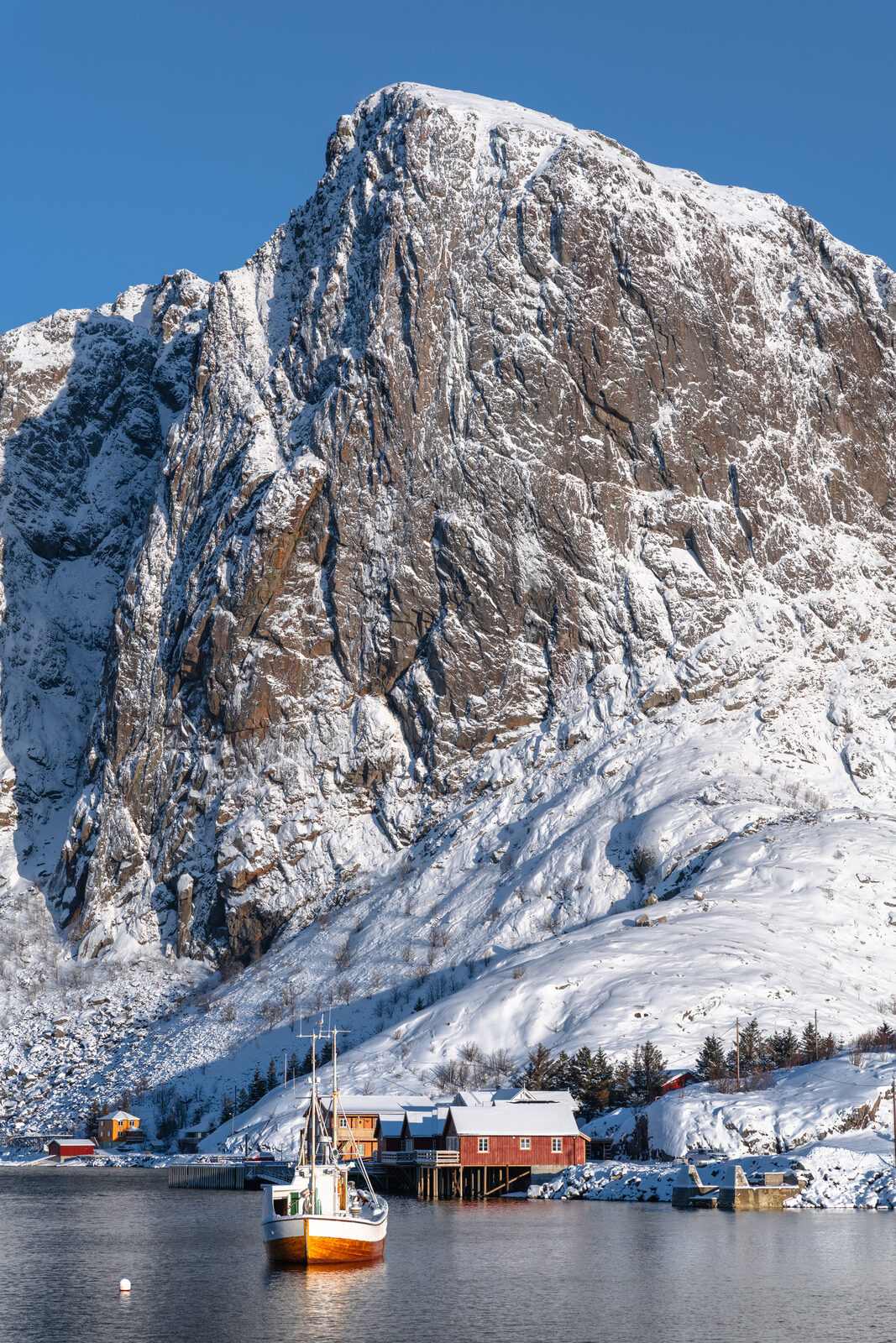 A fishing boat leaving harbour below a snow-covered rock face, red rorbuer on the shore